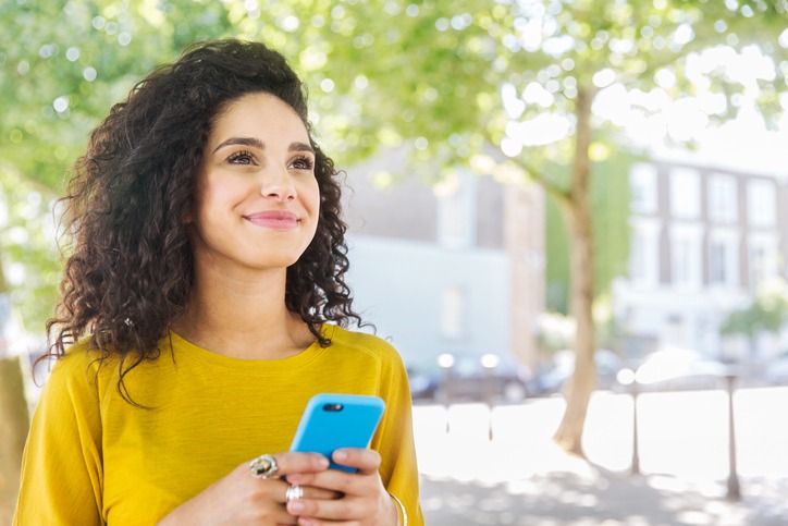  Mulher de cabelos cacheados e blusa amarela olha para o horizonte enquanto usa o celular
