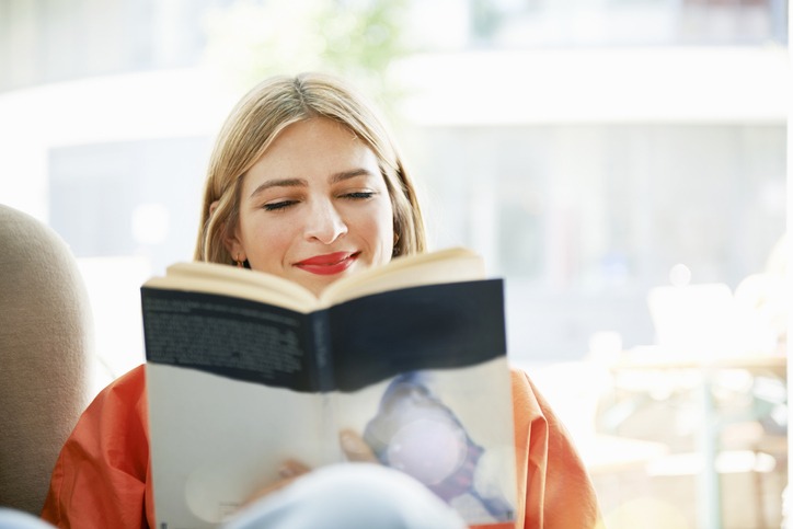 Mulher loira e de batom vermelho lê um livro sentada no sofá
